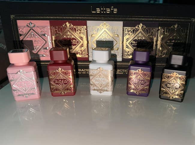 Set of six luxurious perfume bottles with ornate designs, alongside a Nivea articulator balm, displayed connected a reflective surface