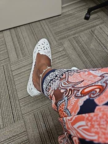 Person wearing achromatic woven flats and patterned pants, seated connected a textured carpeted floor