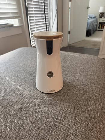 A Furbo canine camera, a cylindrical instrumentality with a wood-like top, sits connected a grey surface, designed for monitoring and interacting with pets remotely