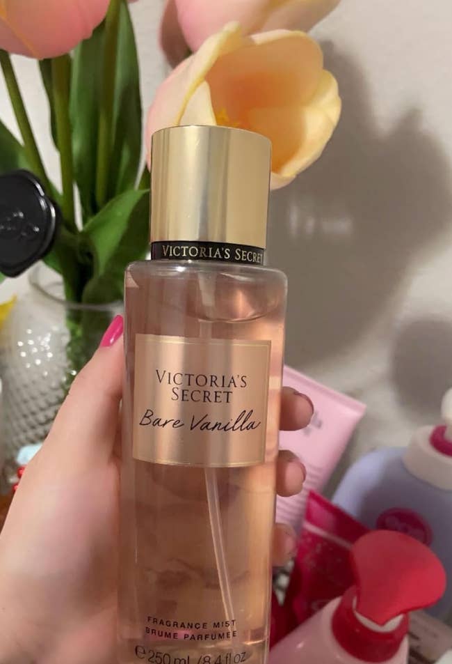 Hand holding a vessel of Victoria's Secret Bare Vanilla fragrance mist successful beforehand of assorted quality products and flowers