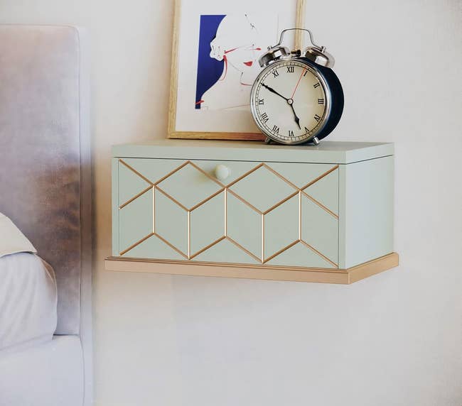 light green floating nightstand on a wall with one drawer and holding a clock and framed art print on top