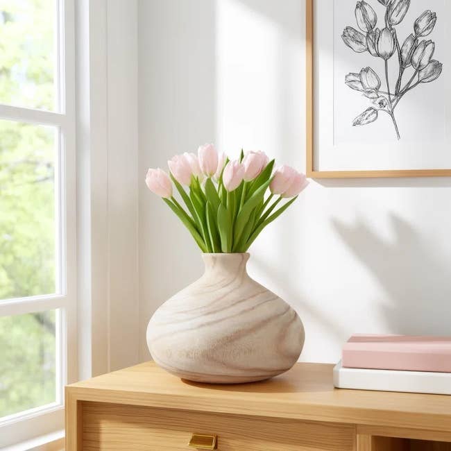 Wooden vase with pink tulips on a wooden table, framed flower sketch on the wall, and books stacked beside in a sunlit room