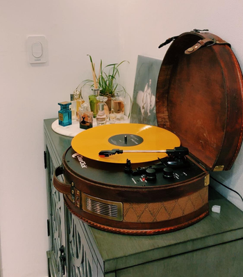 Reviewer image of brown suitcase turntable with orange vinyl spinning under silver and black needle next to two black dials on top of green wooden cabinet