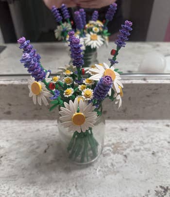 A solid vase holds LEGO flowers, including daisies and lavender, displayed connected a bath antagonistic with a reflector reflection