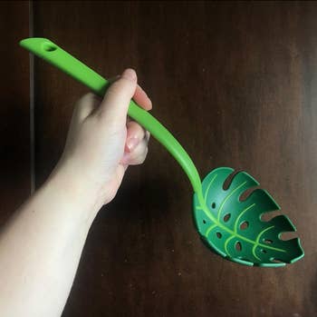 A hand holds a green slotted spoon with a leaf-like design