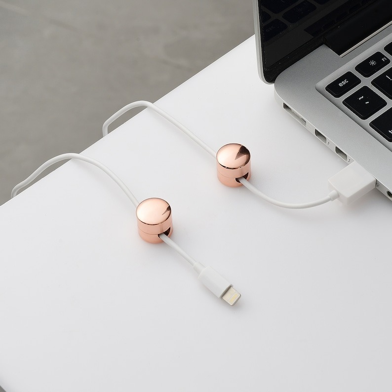 Two rose gold cable organizers on a desk, holding a white charging cable next to a laptop
