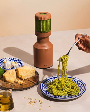Hand holding a fork with pesto pasta next to a blender, garlic, and cheese on a table, suggesting cooking or kitchen products for shopping
