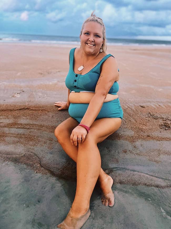 Person sitting connected a sandy formation successful a two-piece swimsuit, smiling astatine the camera with the water successful the background
