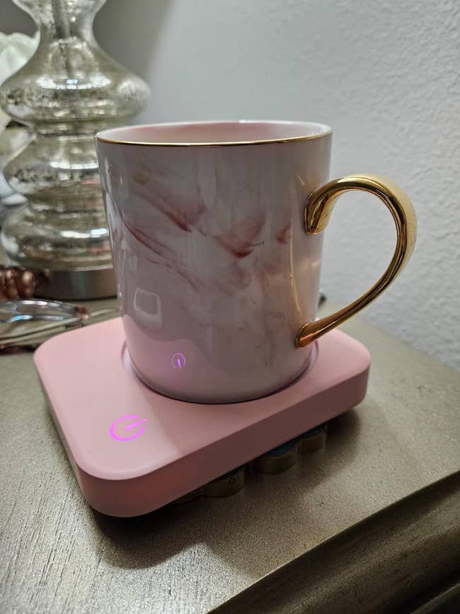 A marble-patterned mug with a metallic handle, on a pink warming base, placed on a nightstand