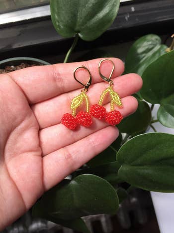 Hand holding cherry-shaped beaded earrings with golden hooks, acceptable against a backdrop of greenish leaves