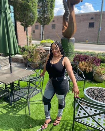 A reviewer stands outside by a patio table in stylish, form-fitting black and gray jeans and a black tank top, smiling at the camera