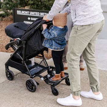 a parent pushing a stroller with the attachment down; a toddler stands on the attachment and holds onto the handle