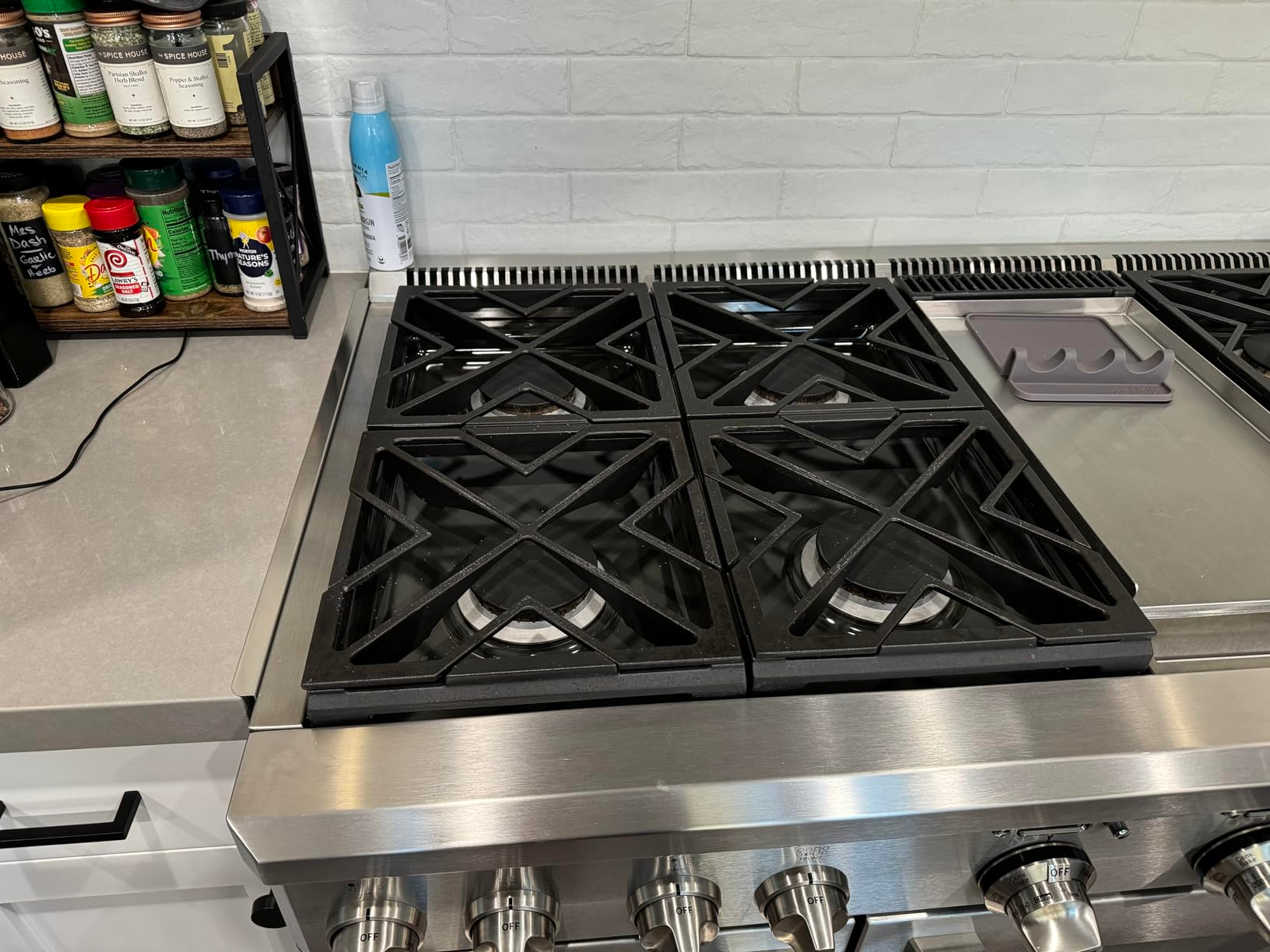 Kompor dapur stainless steel dengan empat pembakar dan beberapa wadah bumbu ditampilkan di rak kayu di dekatnya