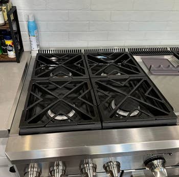 Kompor dapur stainless steel dengan empat pembakar dan beberapa wadah bumbu ditampilkan di rak kayu di dekatnya