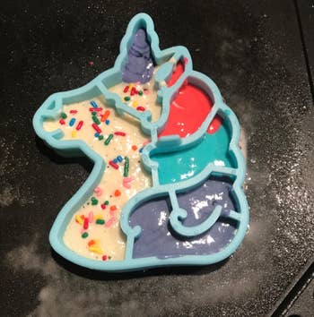 A reviewer's blue unicorn shaped mold on a frying pan with pancake batter in various colors 