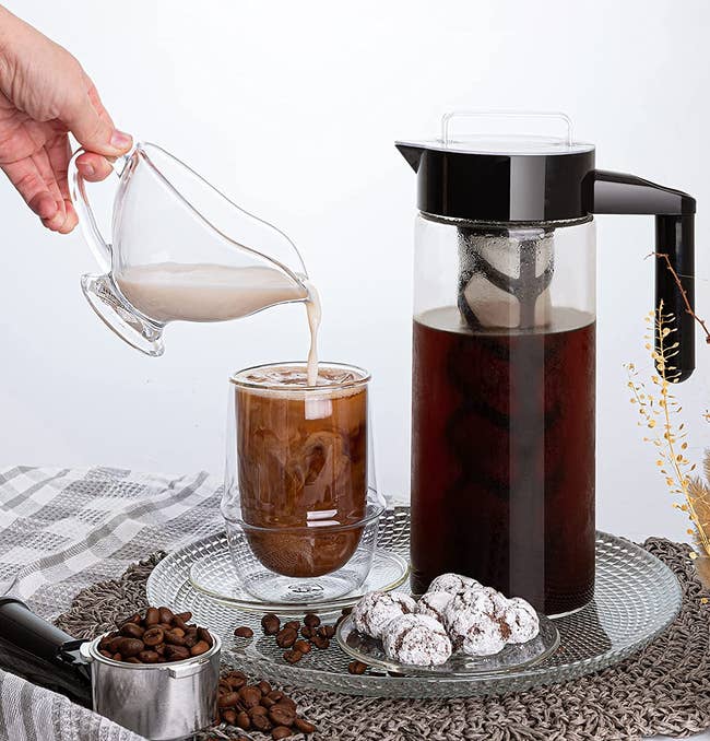 Clear cold brew maker with black handle and filter inside next to model pouring milk into cup of coffee