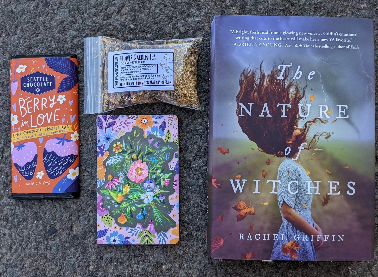 the contents of a subscription box, including a fantasy novel, tea, notebook, and bar of chocolate