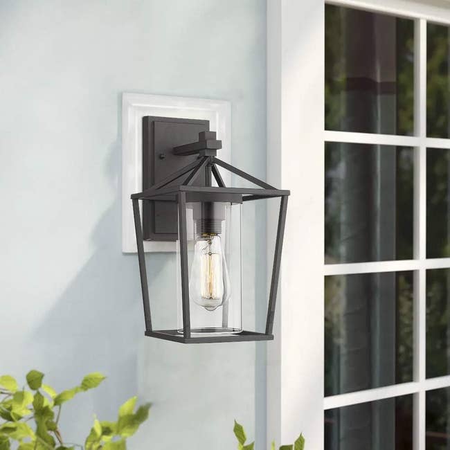 A modern outdoor wall lantern with a geometric metal frame, clear glass panels, and visible filament bulb mounted next to a window