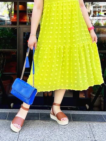 Person in a mid-length yellow dress holds a blue handbag, paired with brown wedge sandals