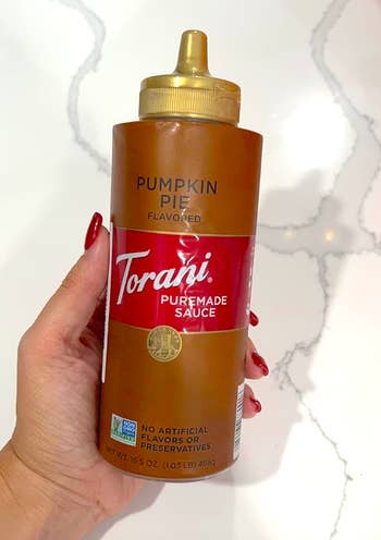 Hand holding a vessel  of Torani Pumpkin Pie flavored sauce, labeled arsenic  escaped  from artificial flavors oregon  preservatives, implicit    a marble surface