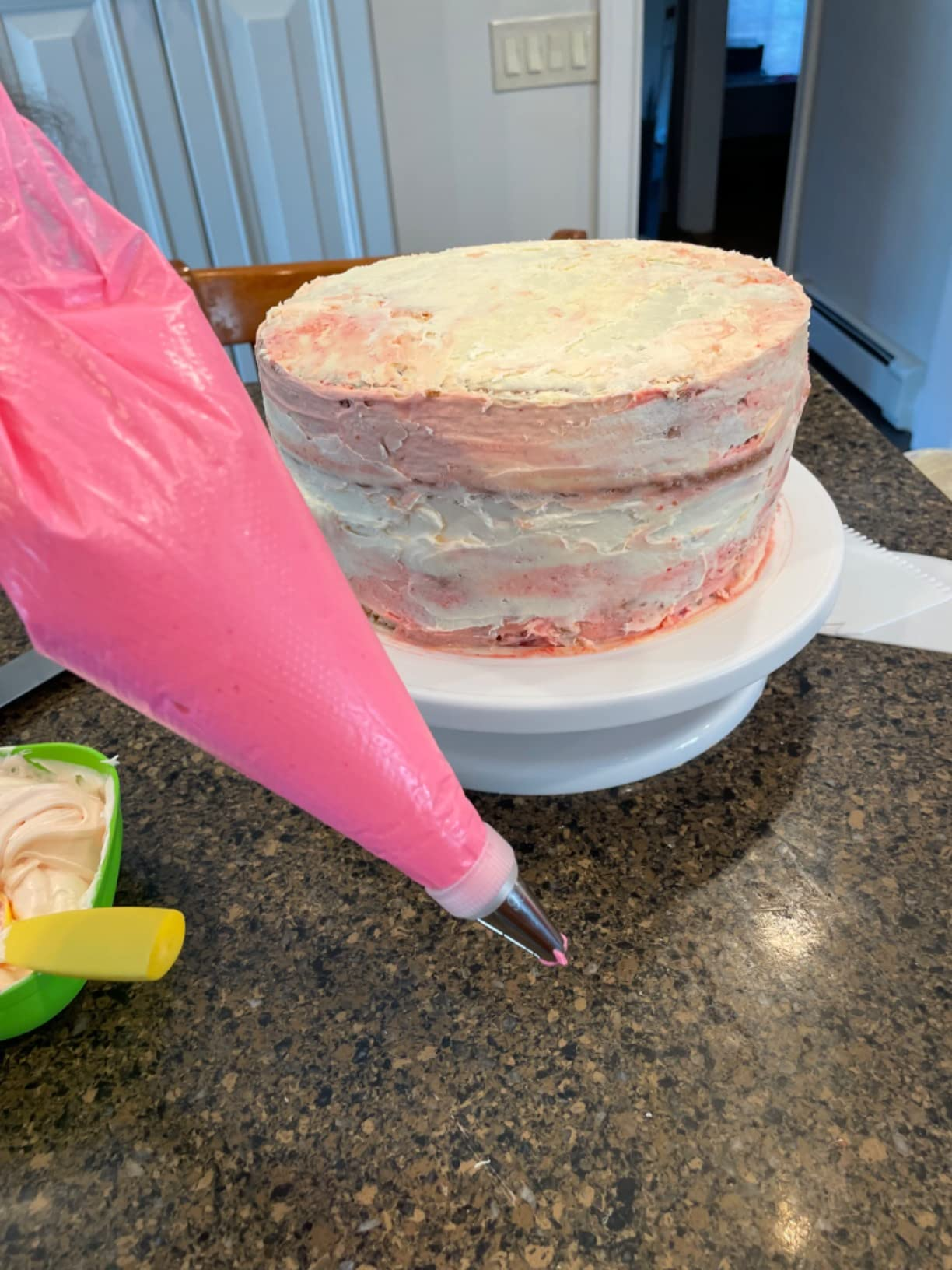 Unfrosted layer cake on a turntable with a piping bag of pink icing ready for decoration on a kitchen counter
