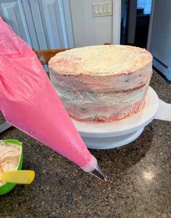 Unfrosted layer cake on a turntable with a piping bag of pink icing ready for decoration on a kitchen counter