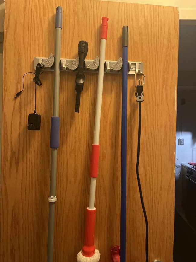Broom and mop organizer mounted connected  a woody  door, holding cleaning tools including a mop, broom, and a plugged-in device