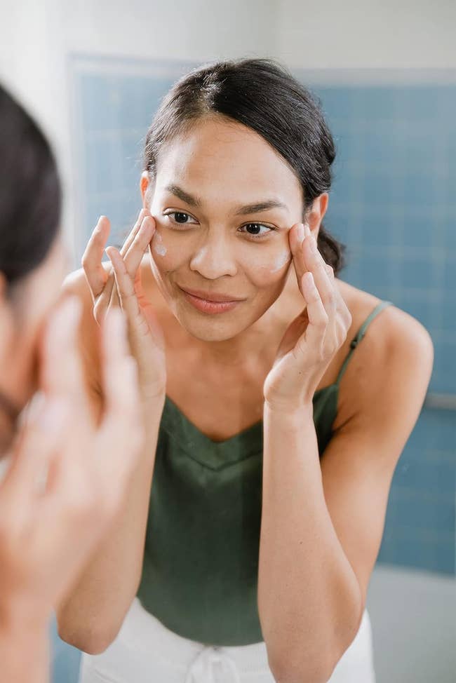 A exemplary  successful  a sleeveless apical  applies pick  to her face, lasting  successful  beforehand   of a mirror, focusing connected  skincare