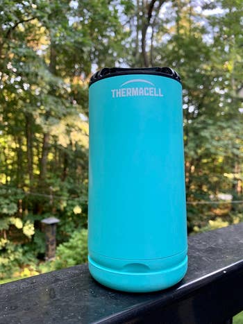Thermacell instrumentality  connected  a railing outdoors, surrounded by trees, utilized  for repelling insects