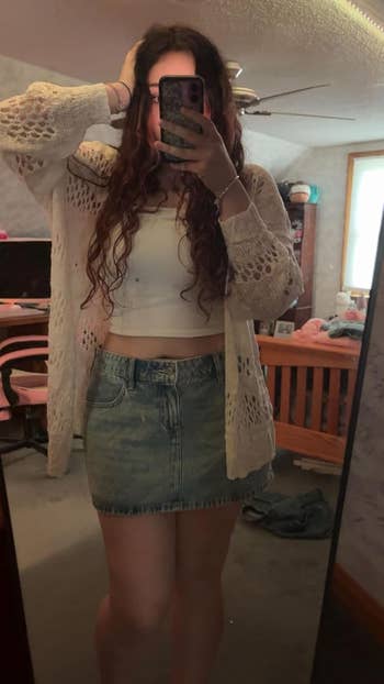Person takes a mirror selfie, wearing a crochet cardigan, white top, and denim skirt in a casual room setting