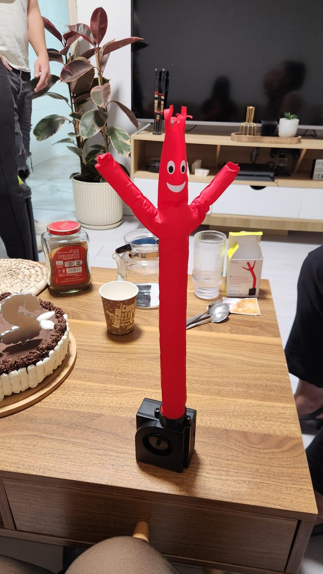 Inflatable red tube man model on a wooden table surrounded by desserts, cups, and decor, suggesting a playful atmosphere