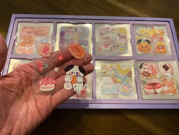 A hand holds animal and food stickers in front of a purple tray filled with more stickers