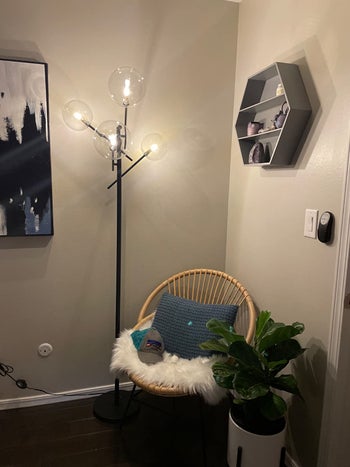 Reviewer image of industrial black lamp with four bulbs in corner of room next to a wicker chair with white cushion and blue throw pillow