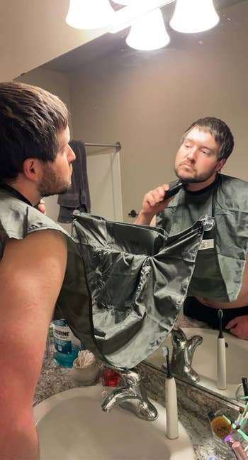 Man using electric trimmer on his beard with a grooming apron in front of a bathroom mirror. Items on sink include a toothbrush and mouthwash