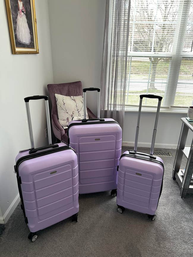 Three purple suitcases of varying sizes with extended handles are displayed in a room with a chair, a painting, and a window