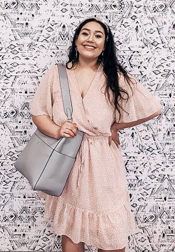Woman in a casual polka-dot dress holding a grey shoulder bag, smiling against a patterned backdrop