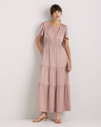 A person models a flowing, floor-length dress with short sleeves and a cinched waist, styled simply with sandals