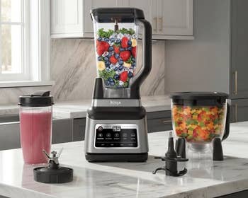 Blender acceptable   connected  a room  antagonistic  with fruits inside, smoothie successful  a to-go cup, chopper with diced vegetables, and blender attachments nearby