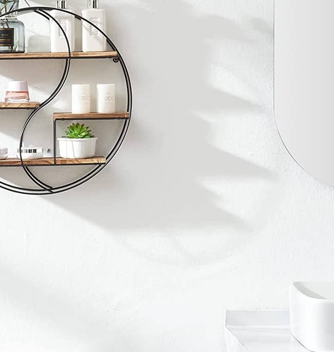 Black metal and wooden circular three-tier shelf mounted to a white wall with toiletries and plants