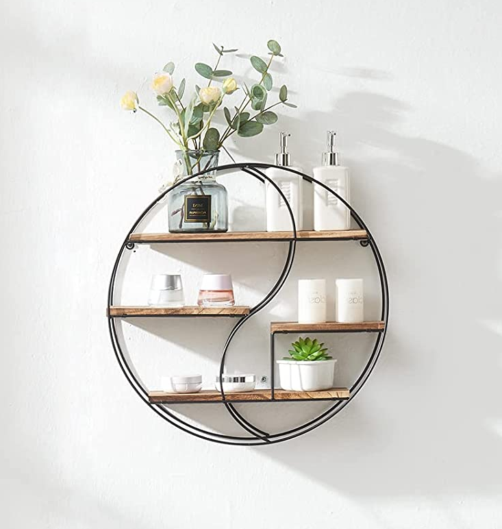 Black metal and wooden circular three-tier shelf mounted to a white wall with toiletries and plants