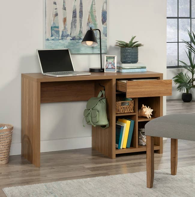 Wooden table with unfastened laptop, lamp, plant, books, photograph frame, and a greenish backpack. A drawer is unfastened showing retention space. Cozy location setup