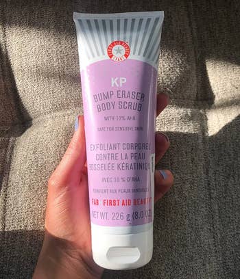 A manus  holds a conduit  of First Aid Beauty's KP Bump Eraser Body Scrub, a merchandise  for delicate  tegument  with 10% AHA, displayed against a textured background