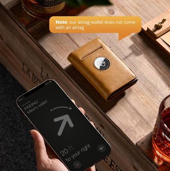 A person holds a phone showing an airtag tracking app. A wallet with an airtag slot is on the table. Note: Airtag not included with wallet