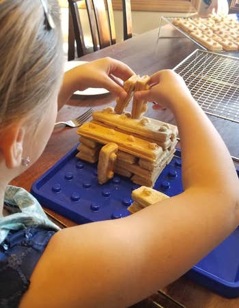 Child building a structure using waffle fries on a blue board, with more fries cooling on a rack in the background