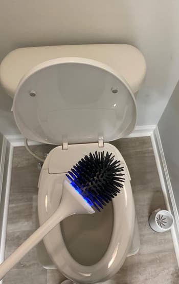 reviewer holding electric silicone toilet brush with blue light on above their open toilet bowl
