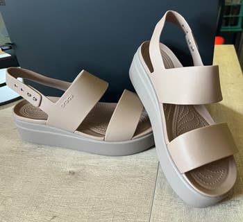 Two beige, level    wedge sandals with wide  straps are displayed connected  a woody  surface