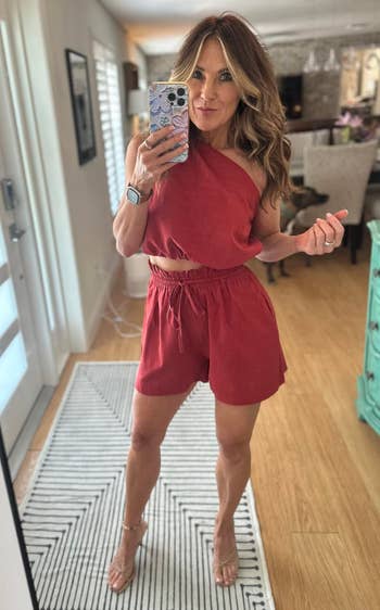 Person taking a selfie in a stylish, casual one-shoulder top and shorts outfit, holding a smartphone