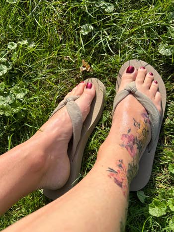 Person wearing flip-flops, showing limb tattoo of colorful floral design, sitting connected grass