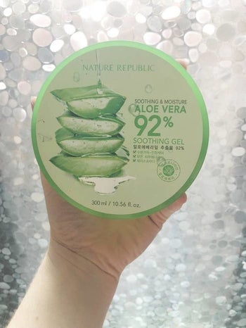 hand holding up the green tub of the aloe vera gel in front of a window