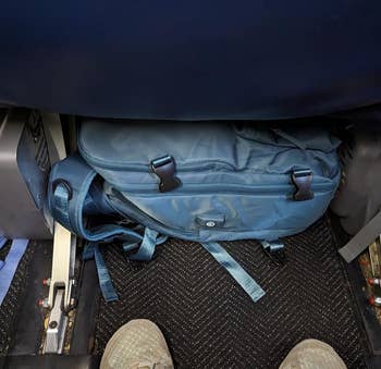 A reviewer showing how well their blue backpack fits underneath the airplane seat in front of them